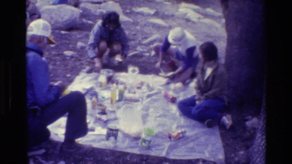 People sit and crouch beside a picnic blanket with food, cans, and bottles in a rocky outdoor setting.
