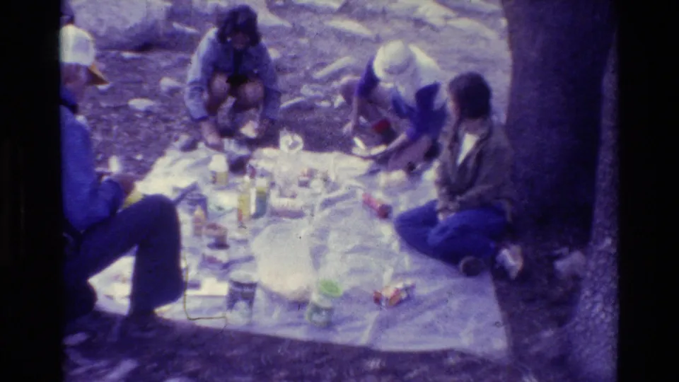 Four visible adults continue arranging a picnic on a white blanket in a rocky outdoor home-movie scene.