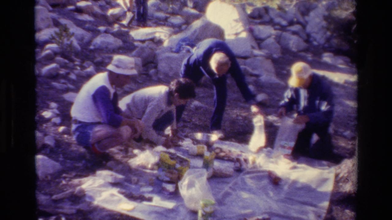 People kneel around a white picnic blanket on rocky ground in bright sun.