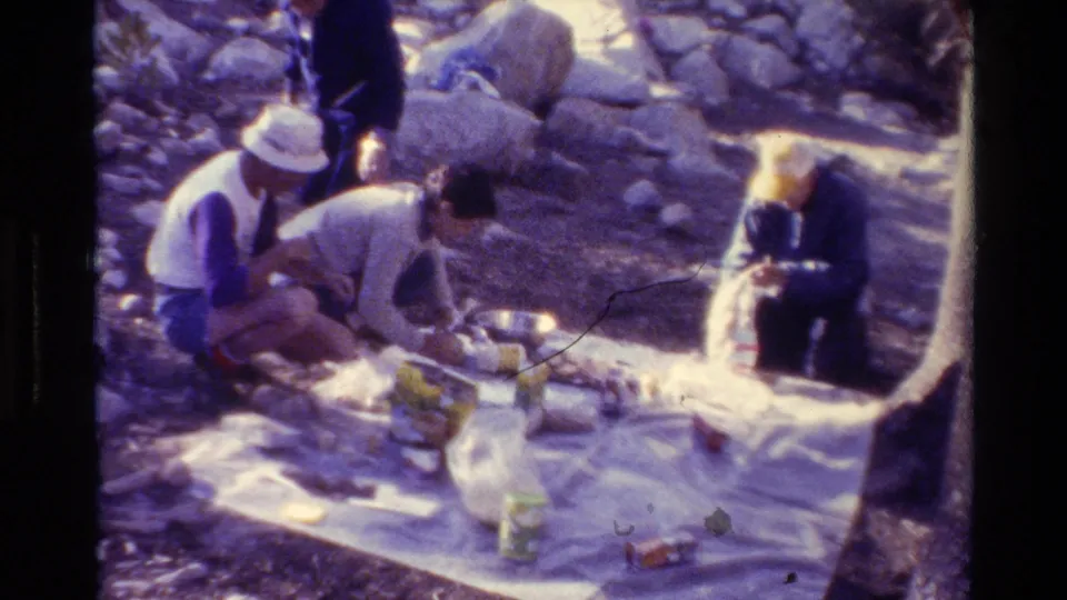 Adults in casual summer clothing arrange food and containers on a blanket during a sunny outdoor picnic setup.