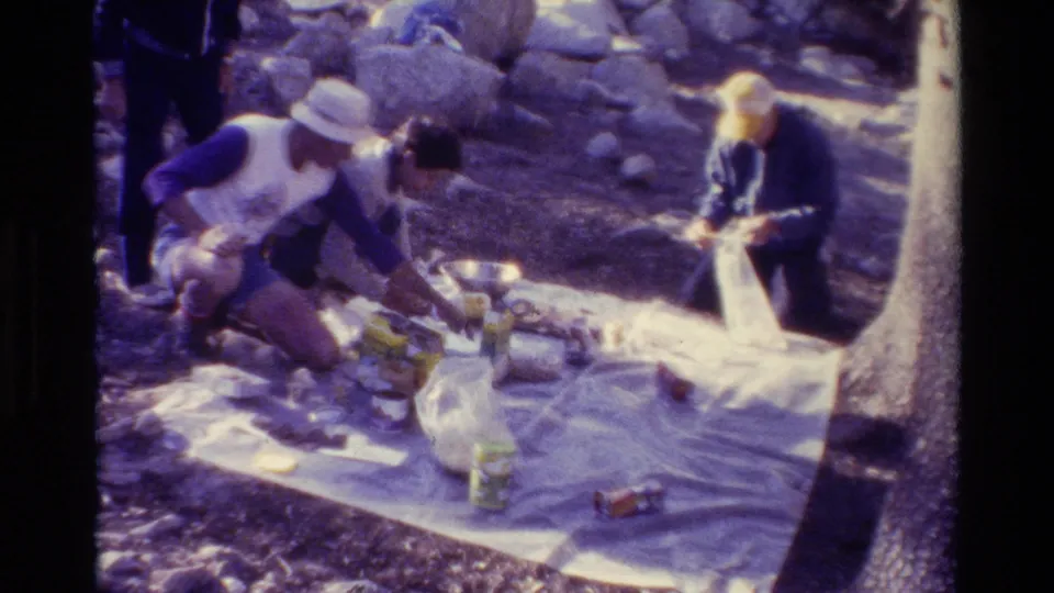 Home-movie view of a small group setting up a picnic spread, with containers and bags scattered across a white blanket.