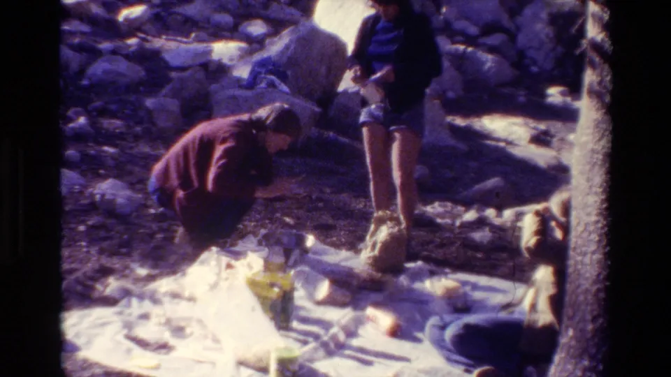 A person in a red top bends over the picnic area as others remain nearby in the rocky sunny outdoor setting.