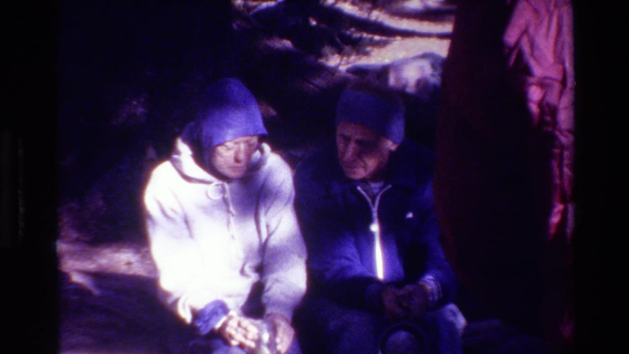 Two adults in blue-toned winter layers sit close together in a snowy forest setting in 1981 home-movie footage.