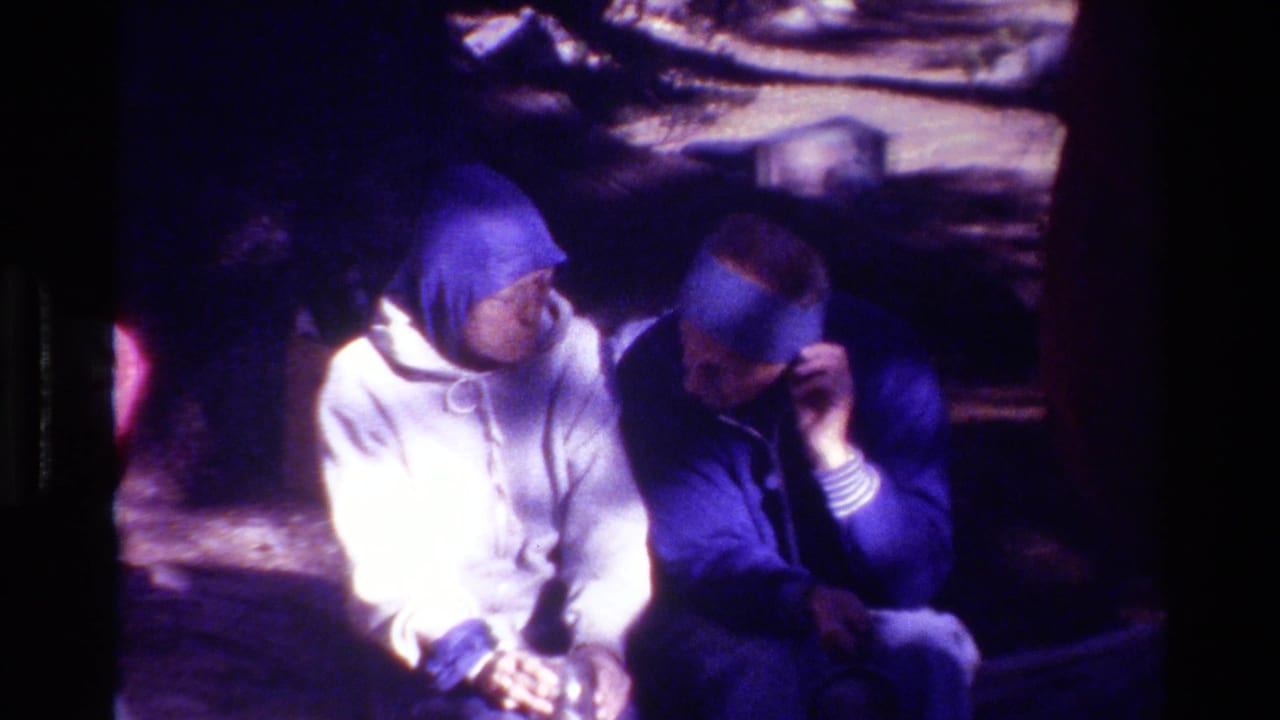 Two adults sit outdoors, one in a white hoodie and one adjusting a blue hat or hood.