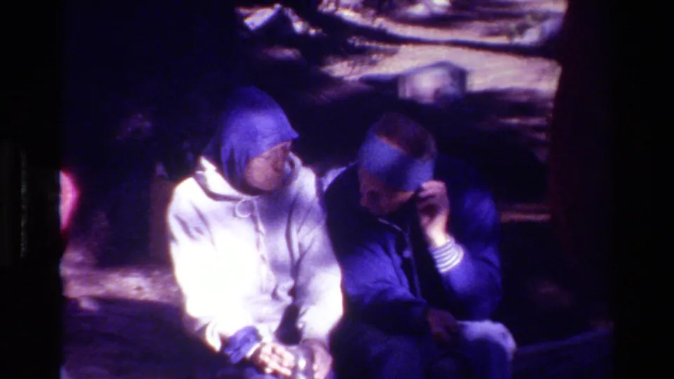 In the 1981 8mm clip, two adults sit in a snowy wooded area while one raises a hand near a blue hat or hood.