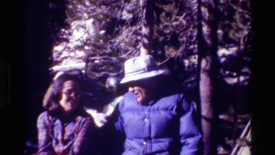 Two adults sit in dappled outdoor light, one smiling in glasses and one wearing a light brimmed hat with a blue jacket.