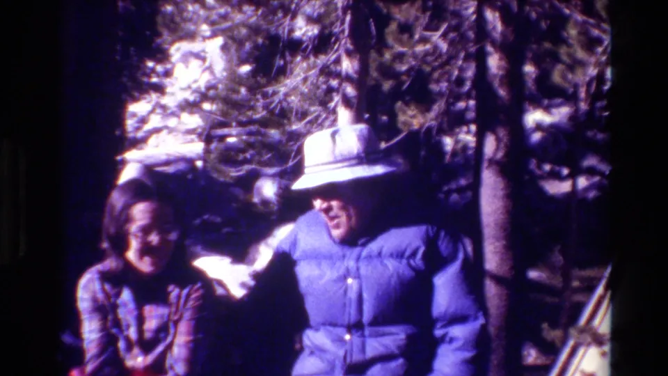 The frame shows seated adults in a wooded snowy setting, with a brimmed hat and blue winter jacket prominent.