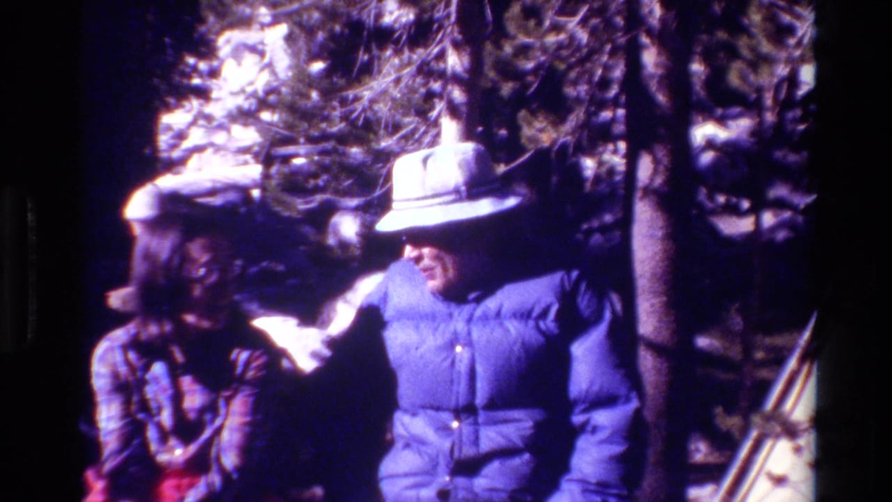 Two adults stand in snowy woods, one in a blue puffer jacket and wide-brimmed hat, with a tent edge nearby.