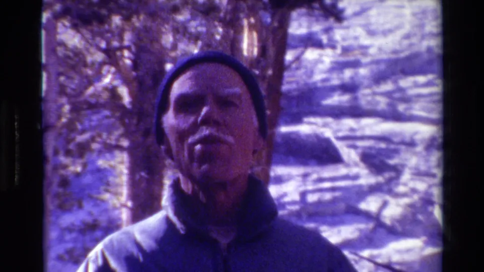 Close 1981 8mm frame of an older man in a dark beanie facing the camera in snowy British Columbia woods.