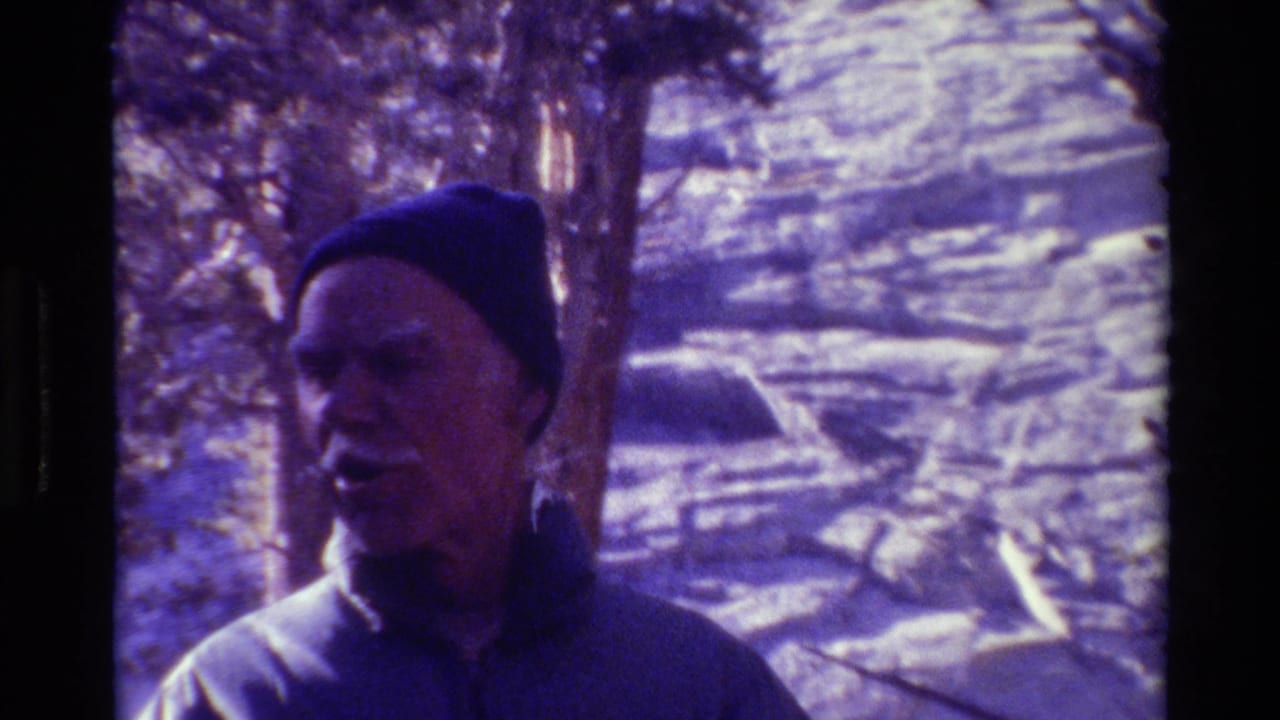 Older man in a dark knit cap standing near trees with bright ground behind him.