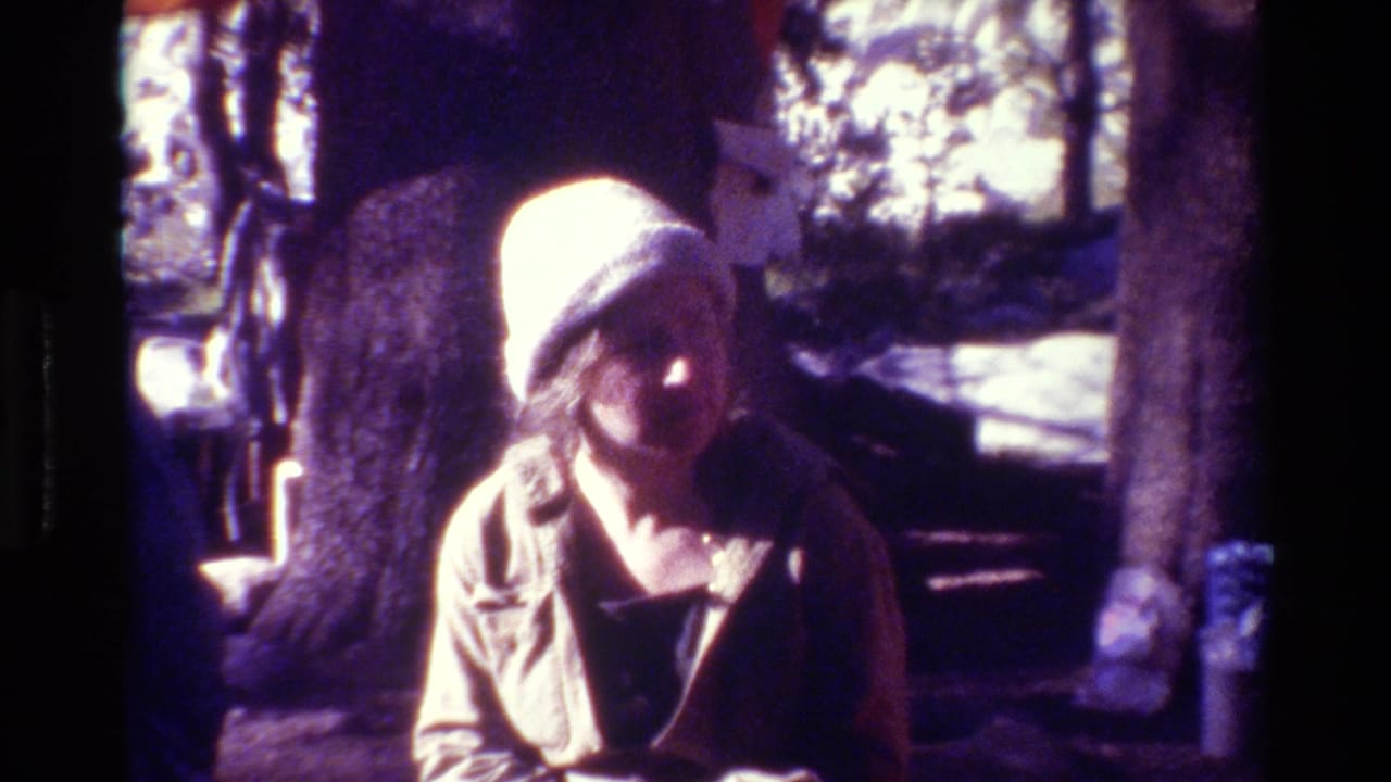 Woman in a light knit hat seated or standing near a large tree in a wooded area.