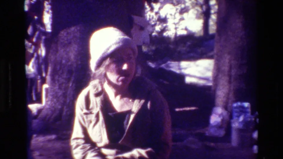 Metadata-supported 1981 8mm frame of a woman in a knit hat looking down in a shaded wooded outdoor scene.