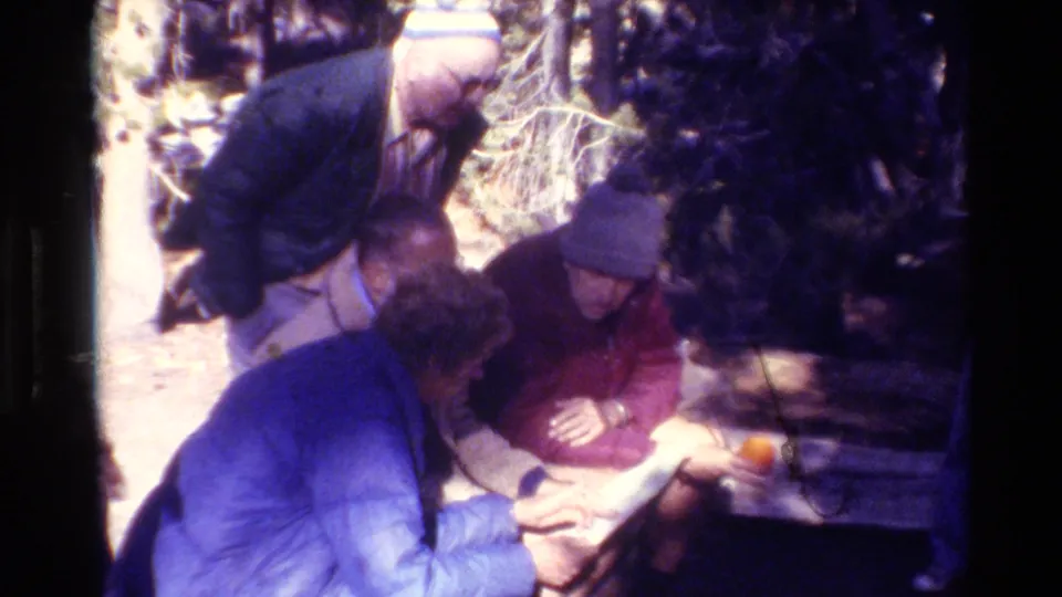A small group of adult men appears to read or discuss papers at a bench in the forest, with strong sunlight and 8mm color shift.