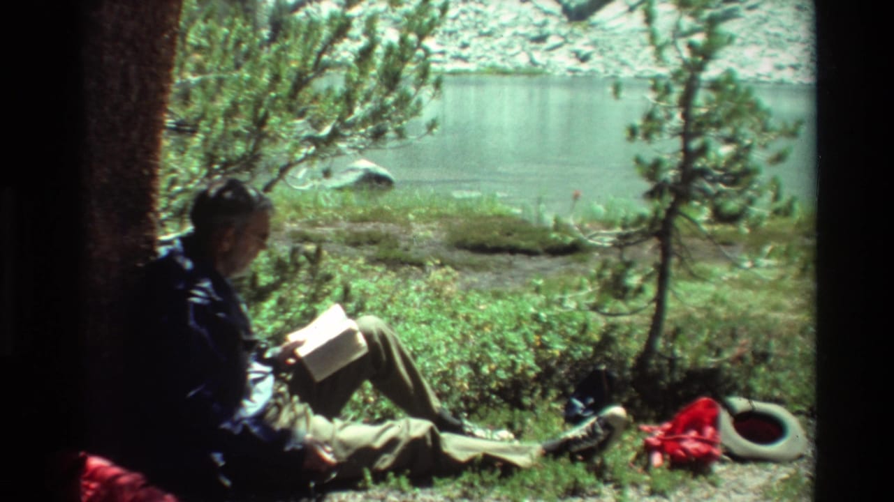 A man in 1981 lakeside home-movie footage sits on grass by calm water, reading beside trees and outdoor gear.