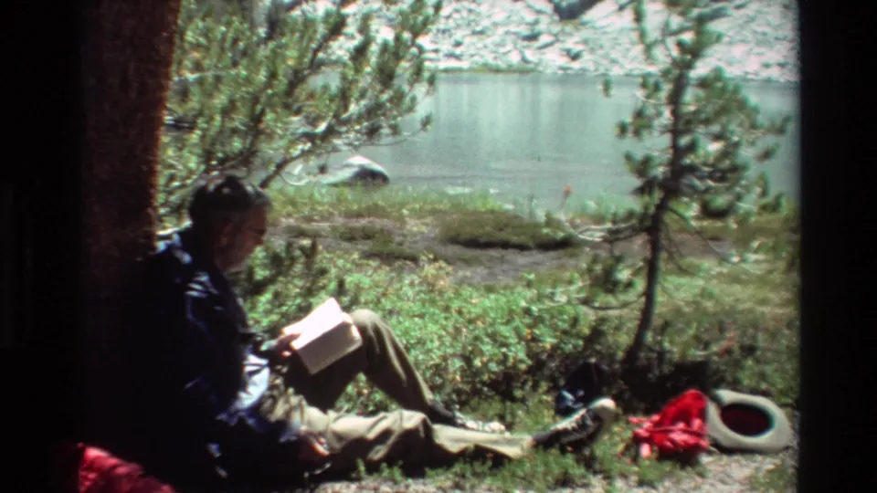 The lakeside reader remains seated in 1981 8mm footage, surrounded by trees, grass, calm water, and loose outdoor gear.