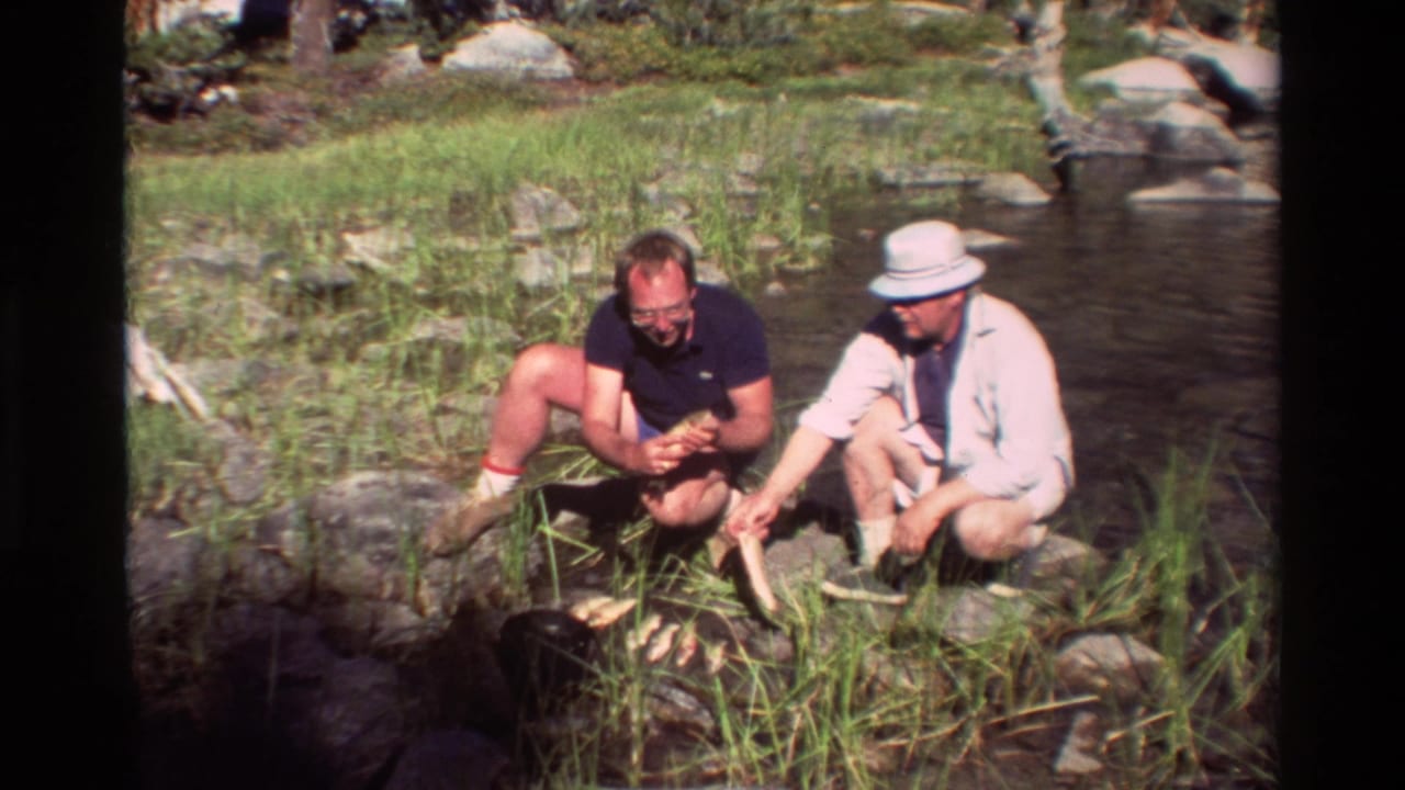 Two adult men crouch beside a rocky stream, with one holding a fish in 1981 8mm home-movie footage.