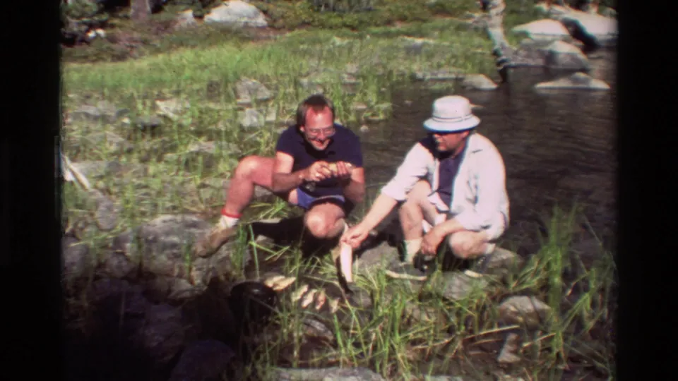 Two adult men crouch in grass near stream rocks, with fish visible by a dark bucket or creel.