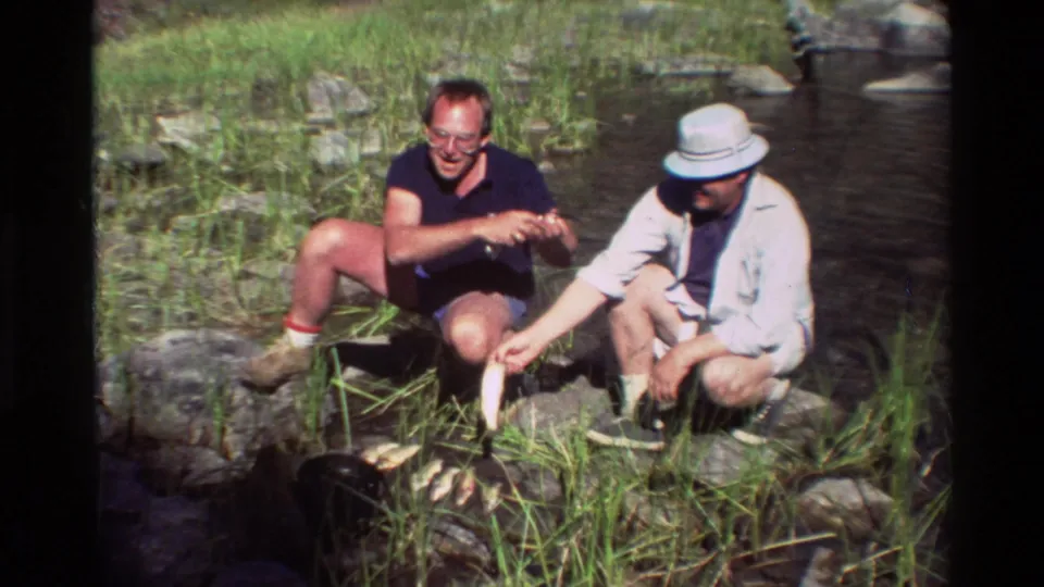 At a rocky stream edge, one adult man in a light hat holds a fish while another man watches nearby.