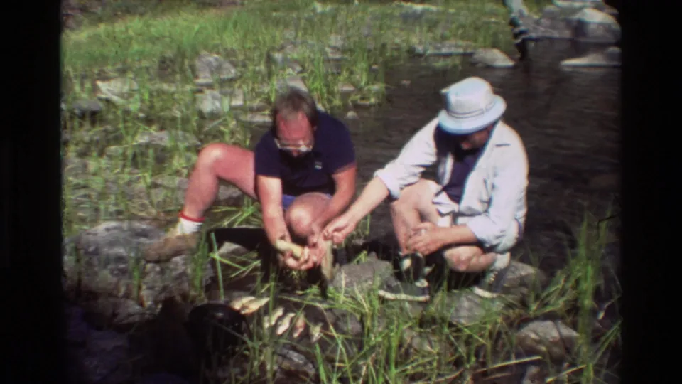 In metadata-supported 1981 British Columbia footage, two adult men crouch beside a shallow rocky stream and handle freshly caught fish.