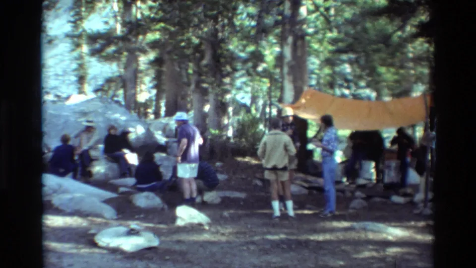 Group of campers in summer clothes stands and sits near a tarp shelter among rocks and pine trees in British Columbia footage.