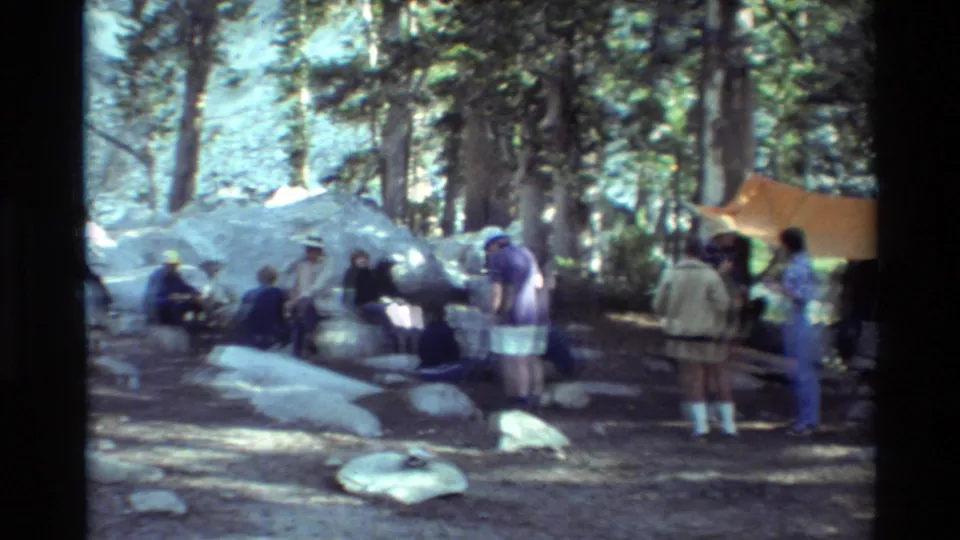 Wide 1981 home-movie view of campers around a forest campsite with rocks, pine trees, and an orange tarp shelter.
