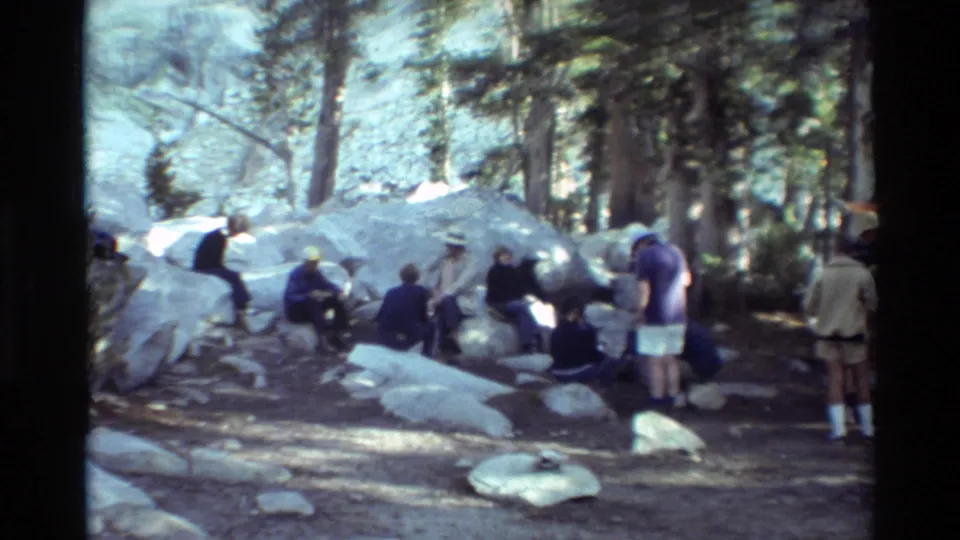 1981 8mm frame of people sitting and standing among large boulders and pine trees in a wooded mountain camping area.