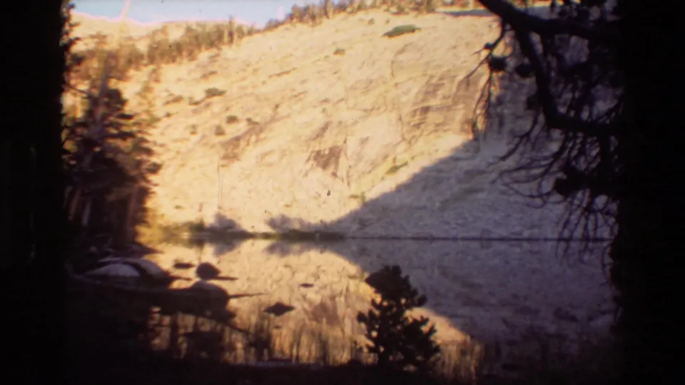 A quiet mountain lake reflects a bright rocky slope and dark tree silhouettes in the 1981 home-movie footage.