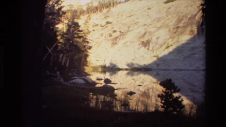 Metadata-supported 1981 British Columbia lake view showing a pale rocky hillside mirrored in calm water beside dark forest edges.