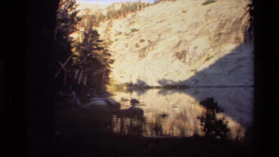 Warm 8mm frame of a quiet mountain lake, with a bright slope, conifers, and shoreline vegetation reflected in still water.