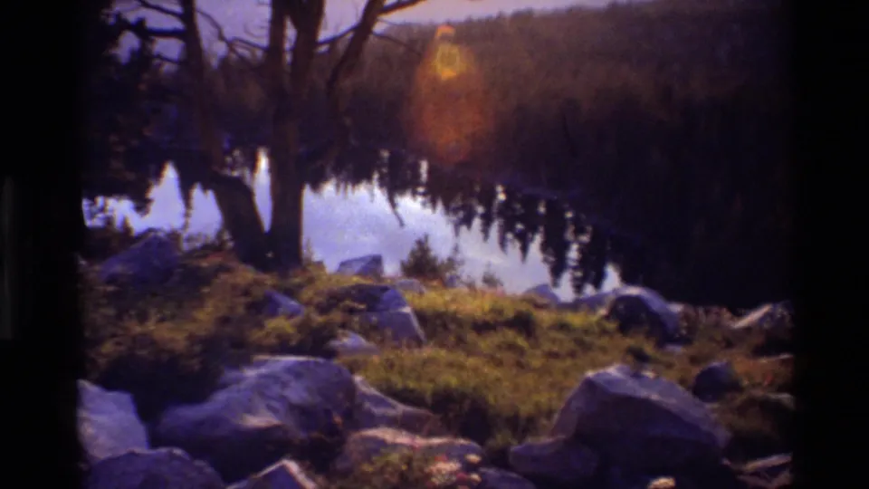 1981 8mm home-movie frame of a rocky lakeshore with bare tree silhouettes, calm water, forest reflection, and distant hills in British Columbia metadata context.