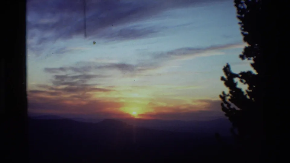 1981 8mm home-movie frame of a sunset over distant ridges, bordered by a dark evergreen silhouette.