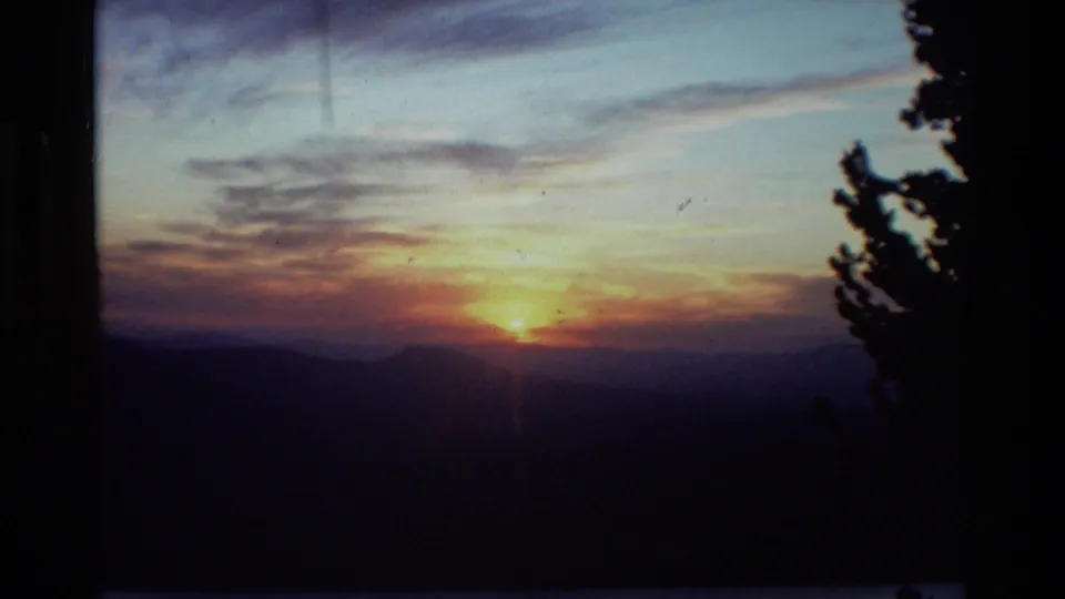 1981 8mm home-movie frame of the sun setting behind dark mountain ridges, with an evergreen silhouette at right.