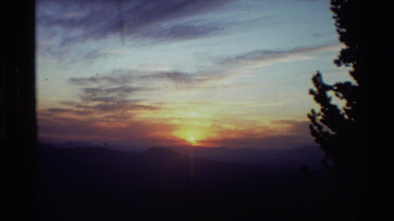 1981 8mm footage of a low sunrise over mountain ridges, framed by dark conifer silhouettes and soft clouds.
