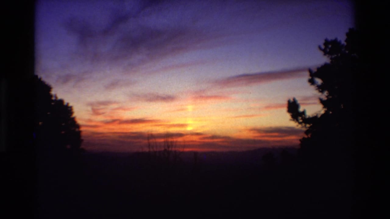 Warm orange sunrise light spreads across clouds over a dark mountain horizon, with trees framing the 8mm shot.