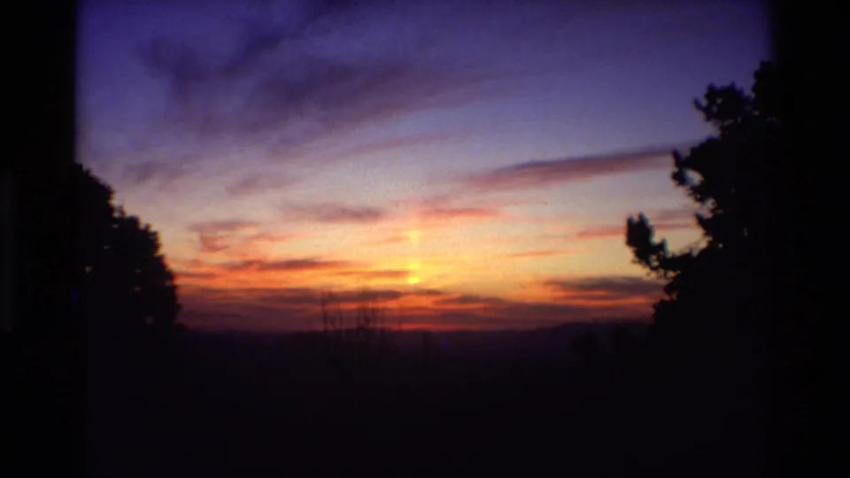 Warm orange sunrise light spreads across clouds over a dark mountain horizon, with trees framing the 8mm shot.