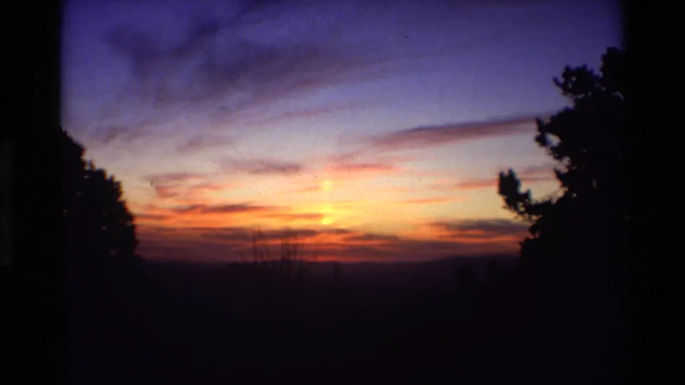 A home-movie view of sunrise clouds over a dark ridge line, with silhouetted trees and visible 8mm frame edges.