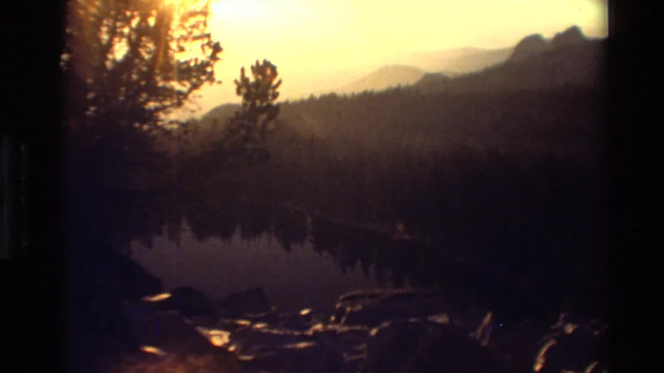 Soft 8mm view of a calm mountain lake at sunrise, with shoreline rocks, forest silhouettes, and hazy ridges.
