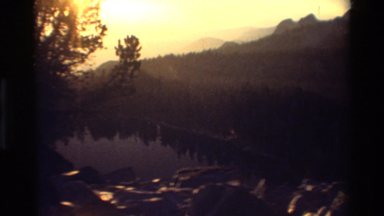 Warm sunset over a dark lake, rocky shore, evergreen trees, and distant mountain ridges.