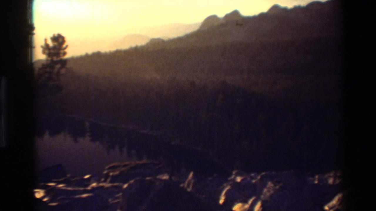 Soft 8mm frame of a forested lake and rocky shore at sunset, with dark mountain ridges in the background.