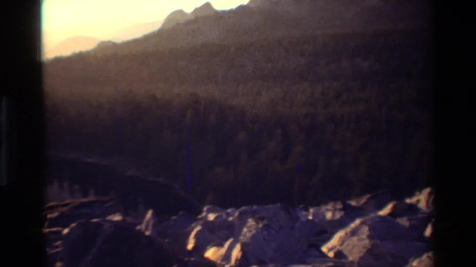 1981 8mm scenic frame looking past foreground rocks and lake water toward dense forest and hazy mountains.