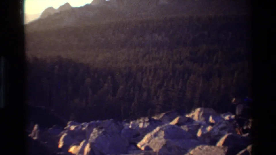 Soft 8mm frame of a rocky lake edge and evergreen-covered slope with a pale mountain ridge behind it.