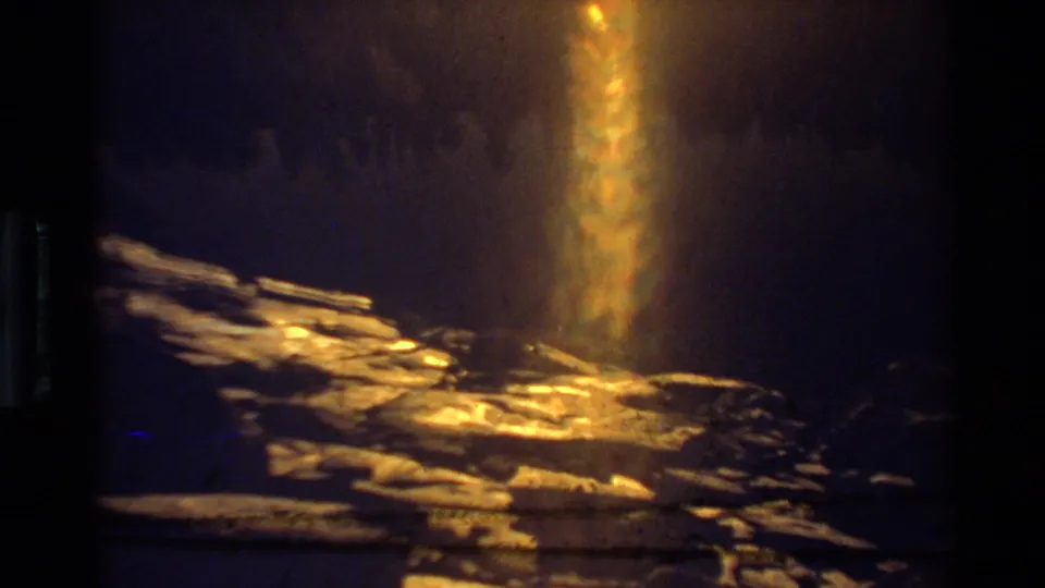 A golden vertical lens flare dominates the frame above rocky ground, with faint shadowed forest texture in the background.