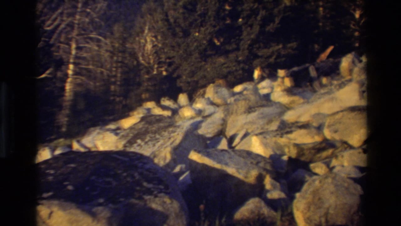 Warm-toned 8mm frame showing boulders, scattered rocks, and trees along a shadowed mountain slope.
