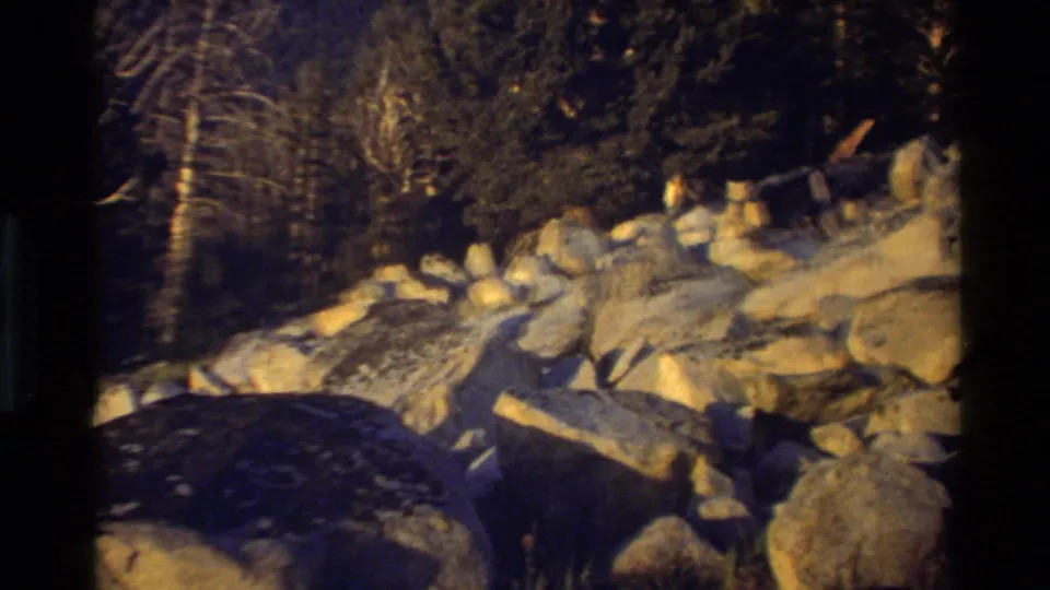 Warm-toned 8mm frame showing boulders, scattered rocks, and trees along a shadowed mountain slope.