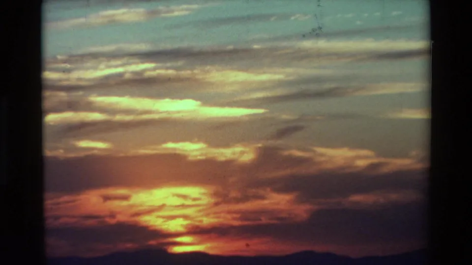 Vintage 8mm frame of a warm sunset sky, pale cloud bands, and a low dark mountain ridge.