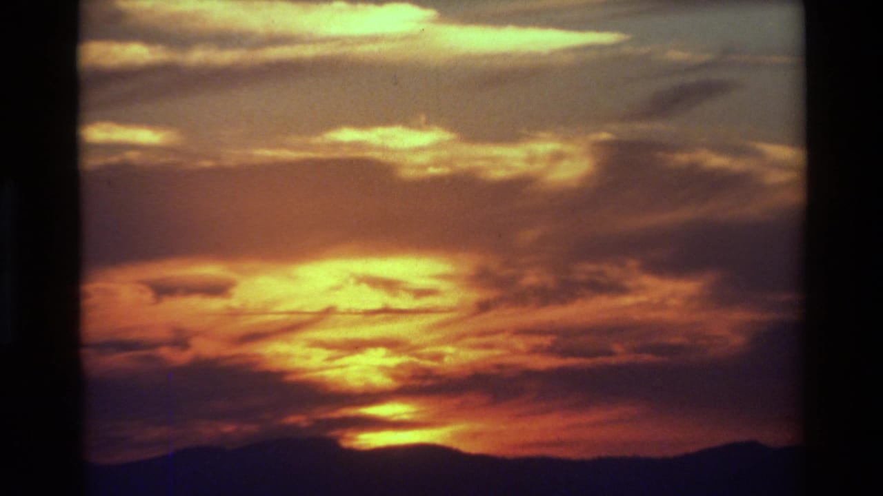1981 8mm footage of a glowing orange sunset above a silhouetted mountain ridge, with layered clouds filling the sky.