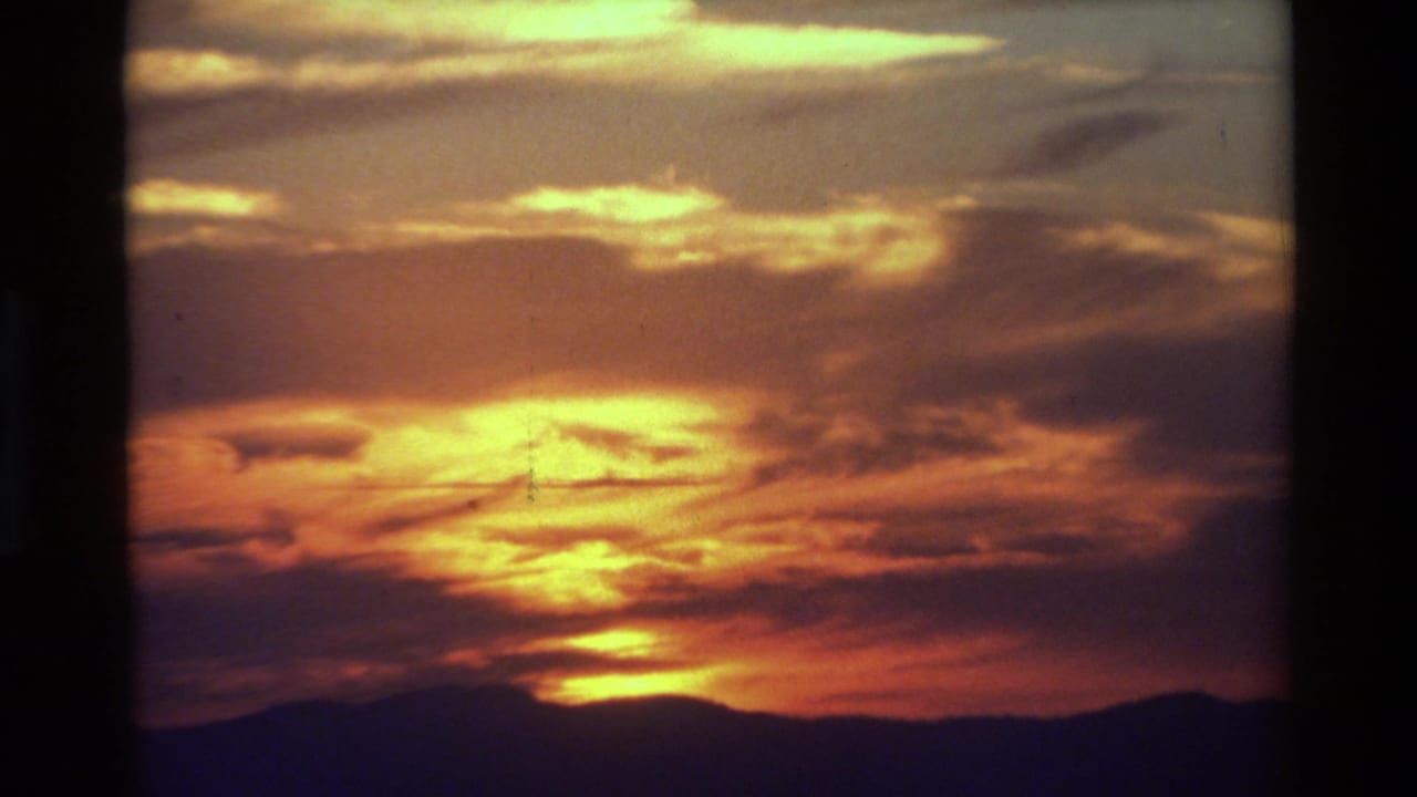 Warm 8mm frame showing bright cloud breaks and an orange sunset over a low mountain silhouette, identified by metadata as British Columbia.