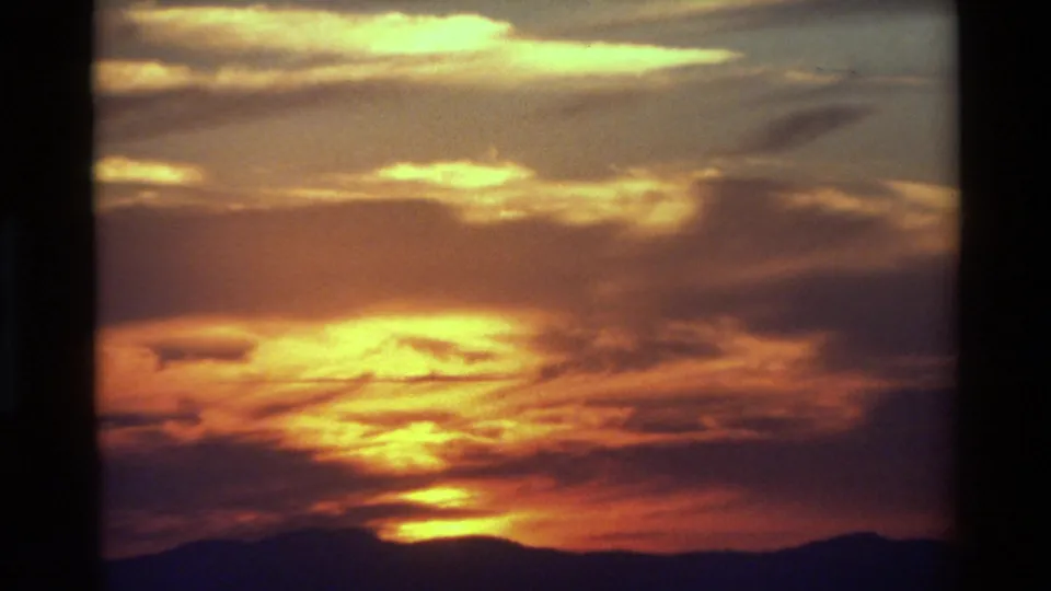 Archival 8mm frame of a saturated sunset sky, with bright yellow cloud bands above a dark, uneven mountain horizon.