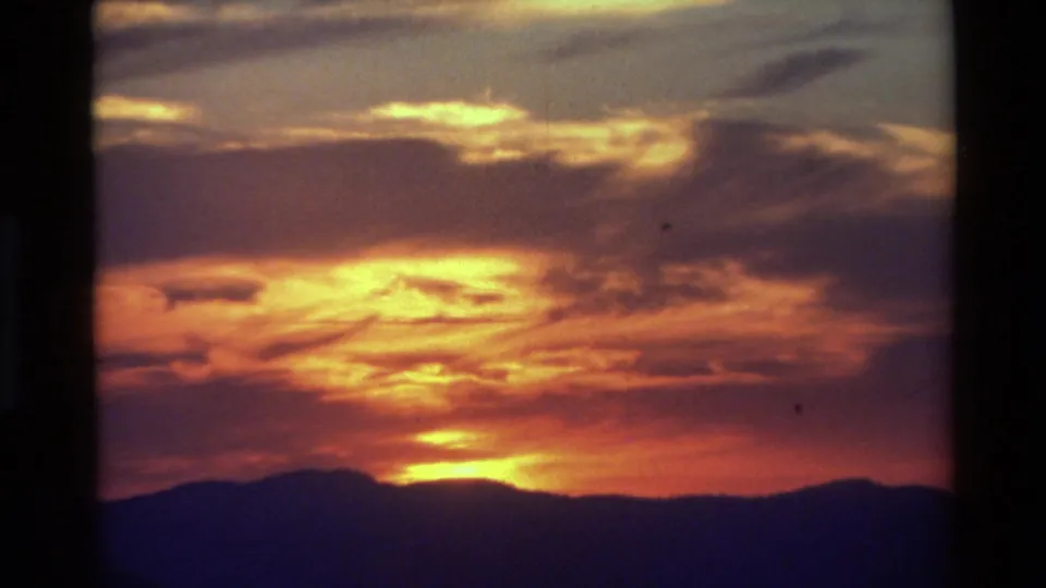 Final 8mm frame showing a deep orange sunset partly hidden by clouds, with the mountain range reduced to a dark silhouette.