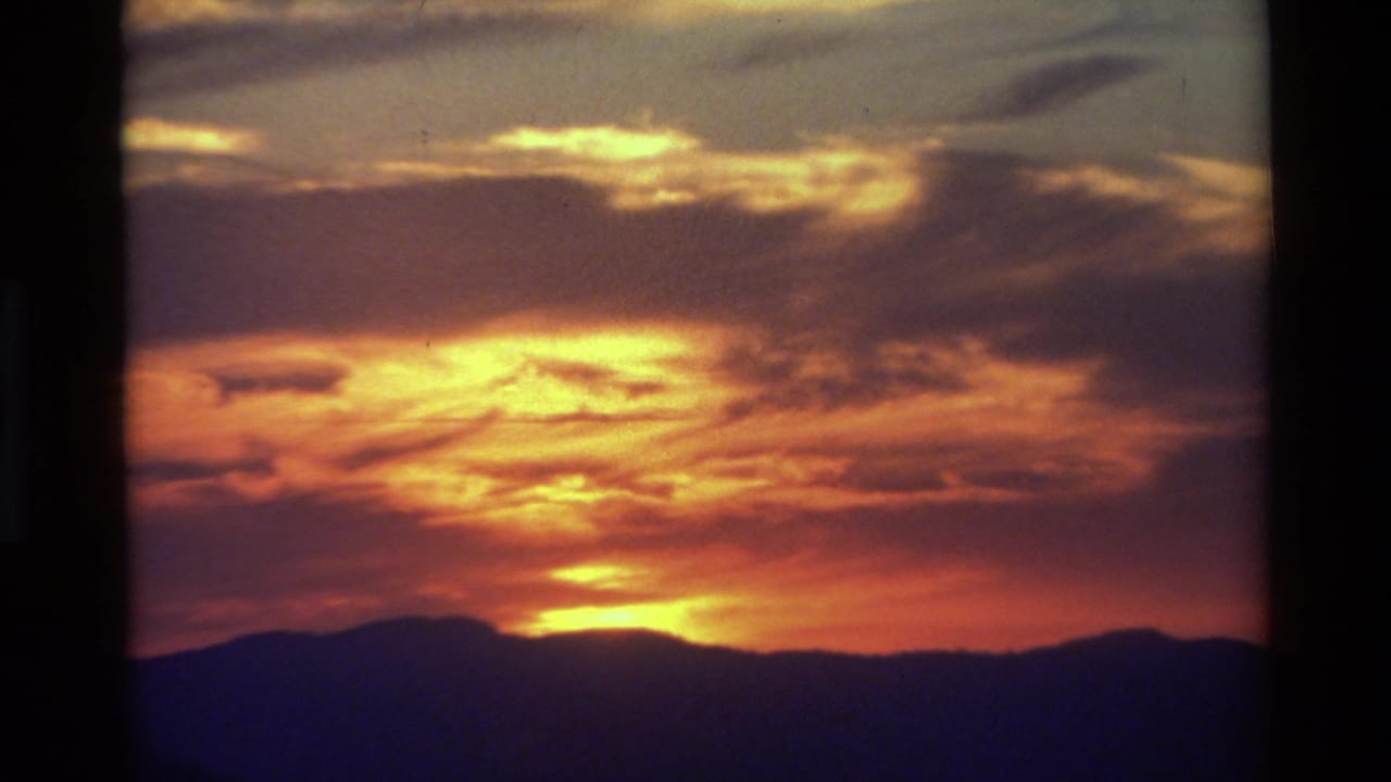 1981 8mm-style view of a warm sunset over silhouetted mountain ridges in British Columbia, with layered clouds above.